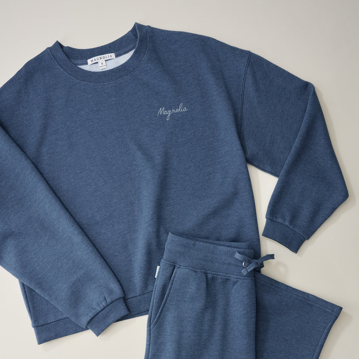 A two-piece kids' loungewear set in heathered blue, featuring a long-sleeve crewneck sweatshirt with 'Maya'sha' embroidered on the chest and matching drawstring sweatpants, laid flat on a light beige background.