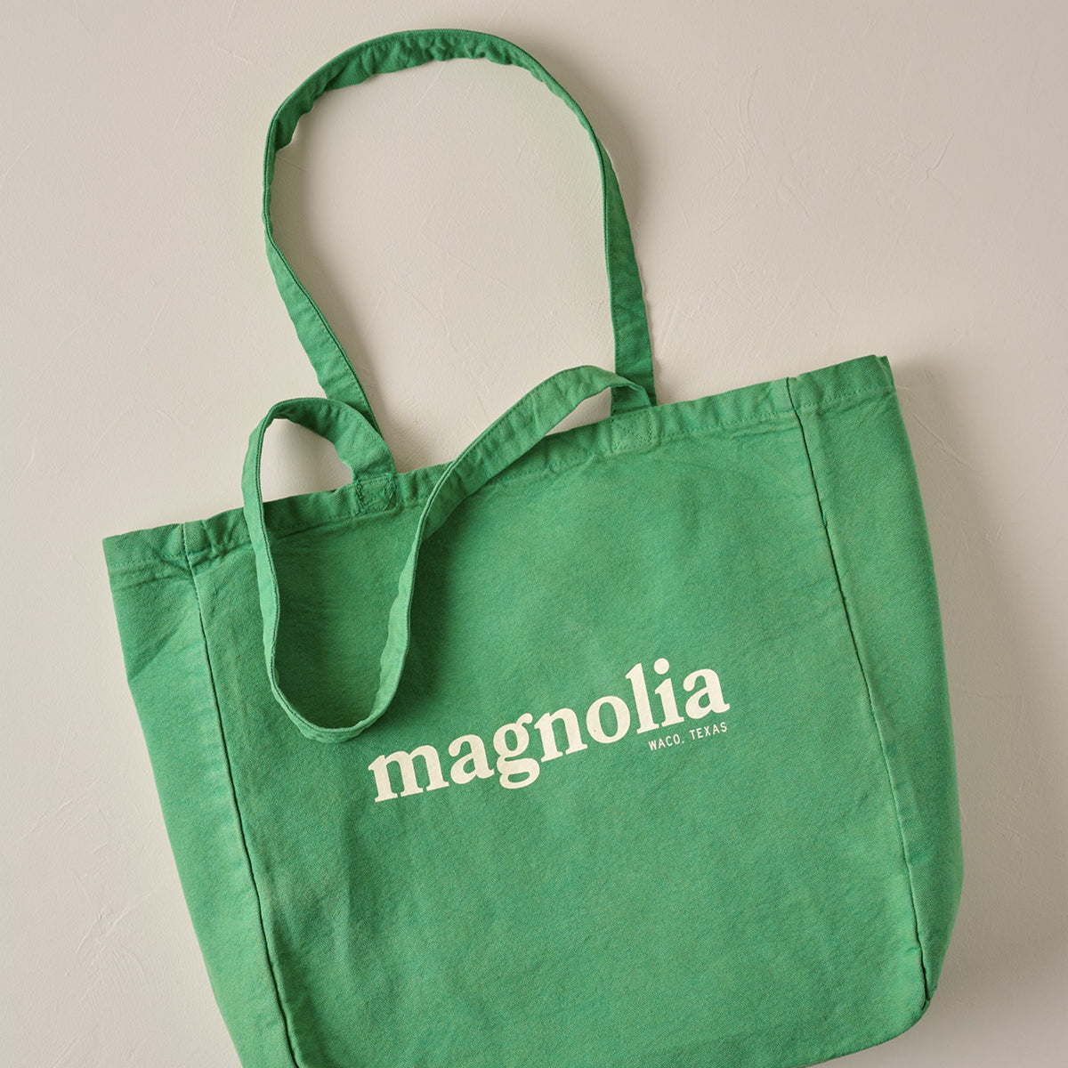 A green canvas tote bag with white printed text reading 'magnolia' and smaller 'NOLA, LA.' on the front, shown against a plain light beige wall.