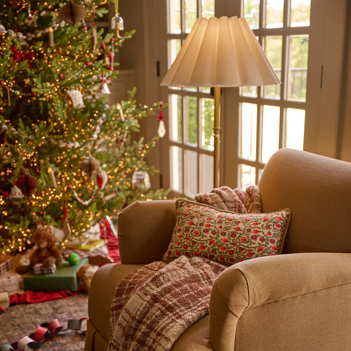 A cozy living room scene with a beige armchair, patterned throw pillow, and plaid blanket, next to a decorated Christmas tree and warm window light.