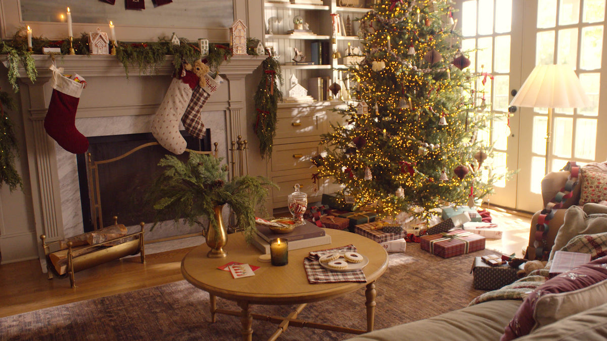 A cozy living room decorated for Christmas, featuring a lit Christmas tree, fireplace with stockings, wrapped gifts, and a round wooden coffee table in the foreground.
