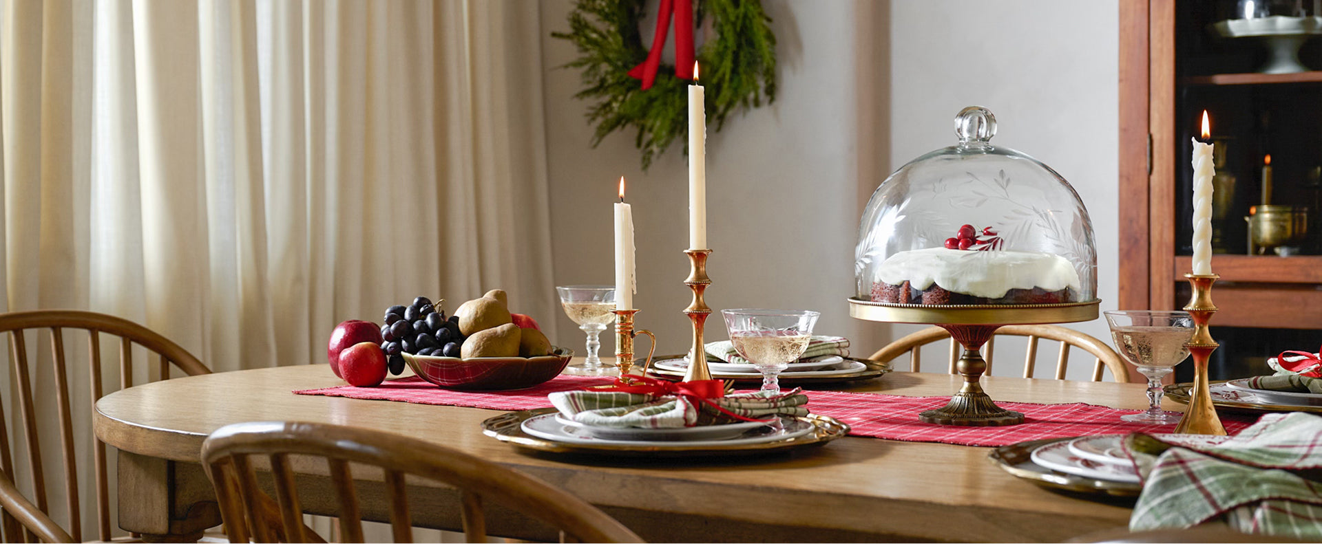 Dining table set for a meal with fruits, candles, and a cake under a glass dome.
