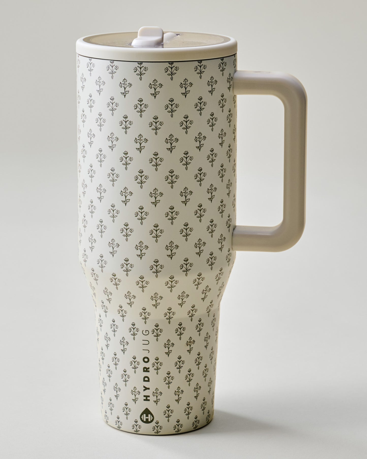 A tall travel mug with a repeating dark gray floral pattern on a warm cream background, featuring a matching cream handle and lid, with the brand name 'HYDRO JUG' printed near the bottom.