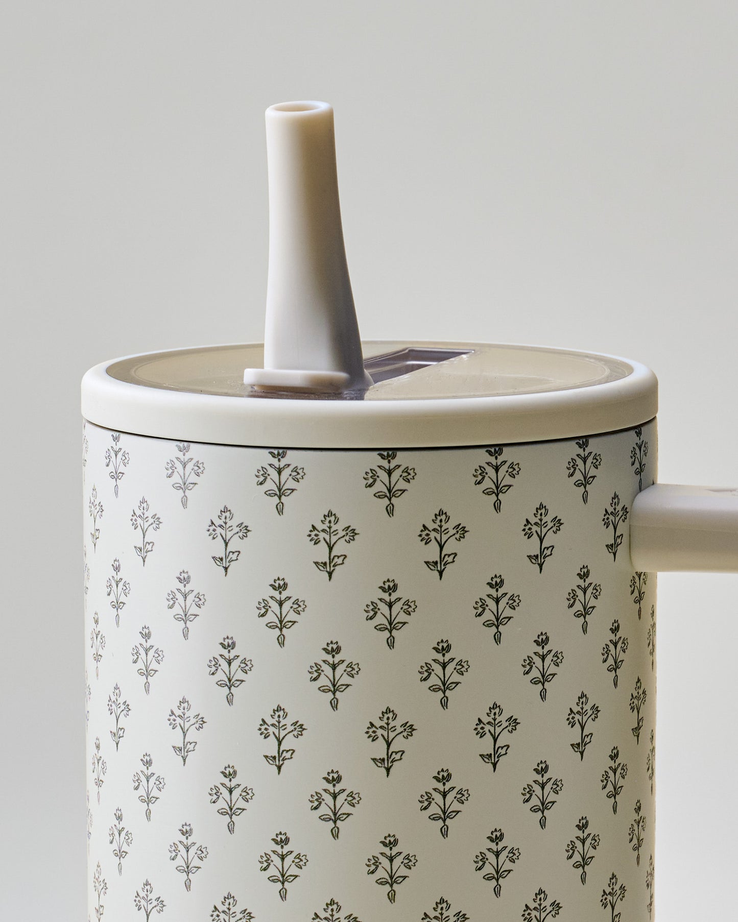Close-up of a cylindrical container with a repeating black floral pattern on a warm off-white background, featuring a light beige lid and spout, photographed against a neutral light gray background.