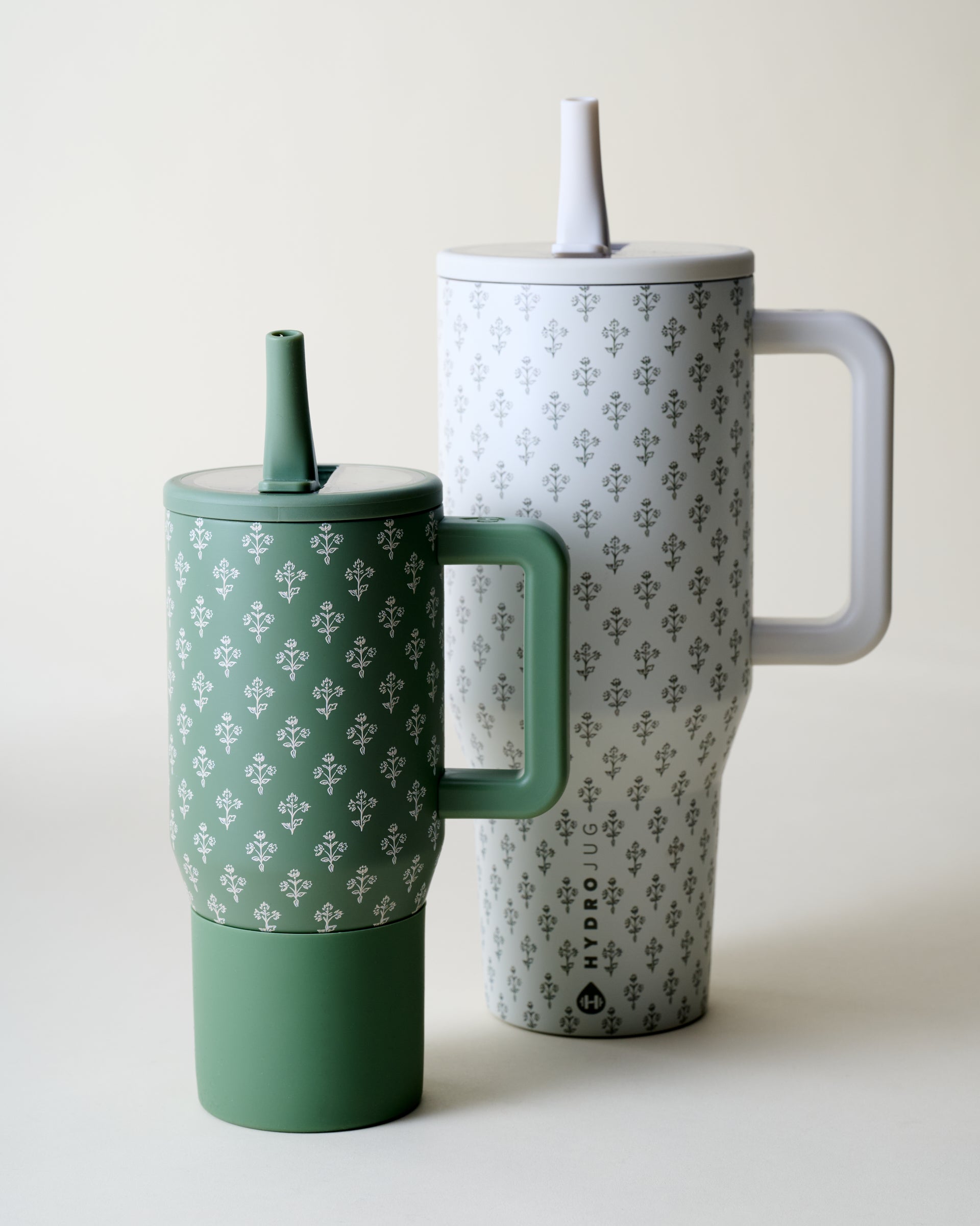 Two HydroJug tumbler cups with lids and handles on a clean off-white background. The cup in front is sage green with a white repeating pattern, and the cup behind is white with a gray repeating pattern.