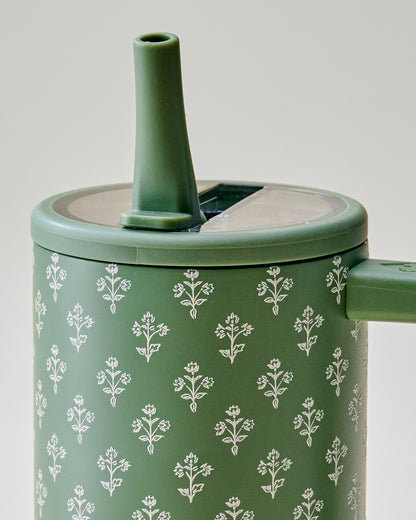 Close-up of a green insulated container or tumbler with a matching lid and spout, decorated with a repeating white floral pattern, on a light neutral background.