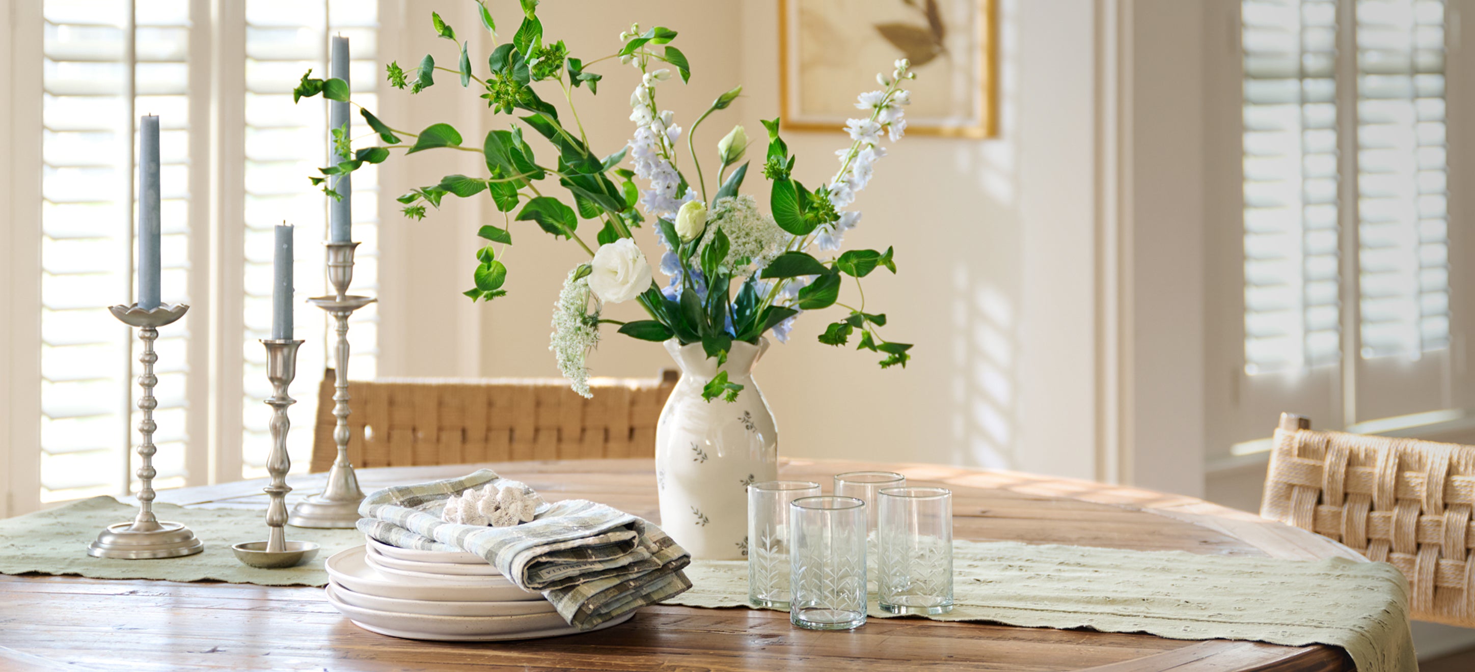 A dining table with taper holders, a stack of dished a white floral vase and drinking glasses on top a green table runner.