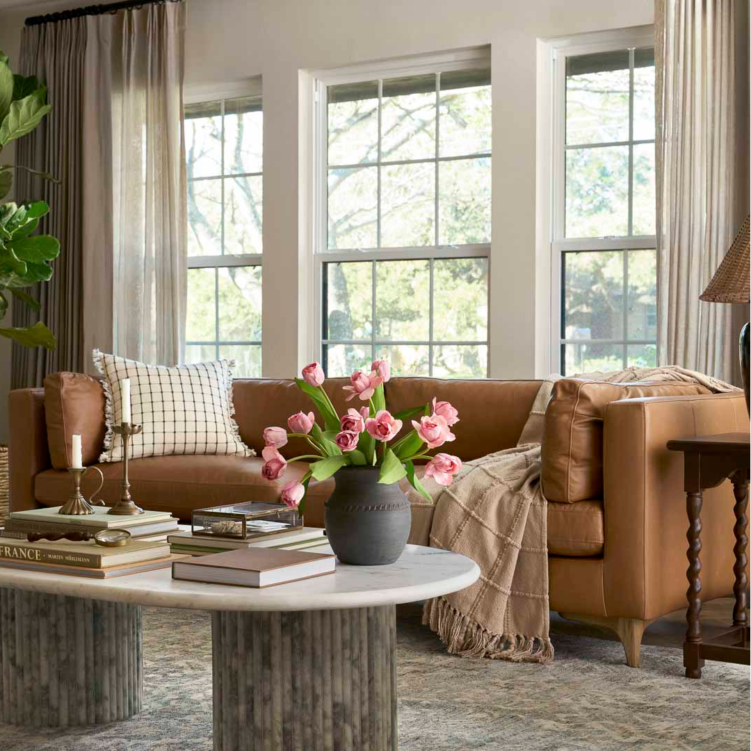 A warm brown leather sofa in a bright, modern living room with tall windows, beige curtains, a round marble-top coffee table, and a vase of pink tulips.