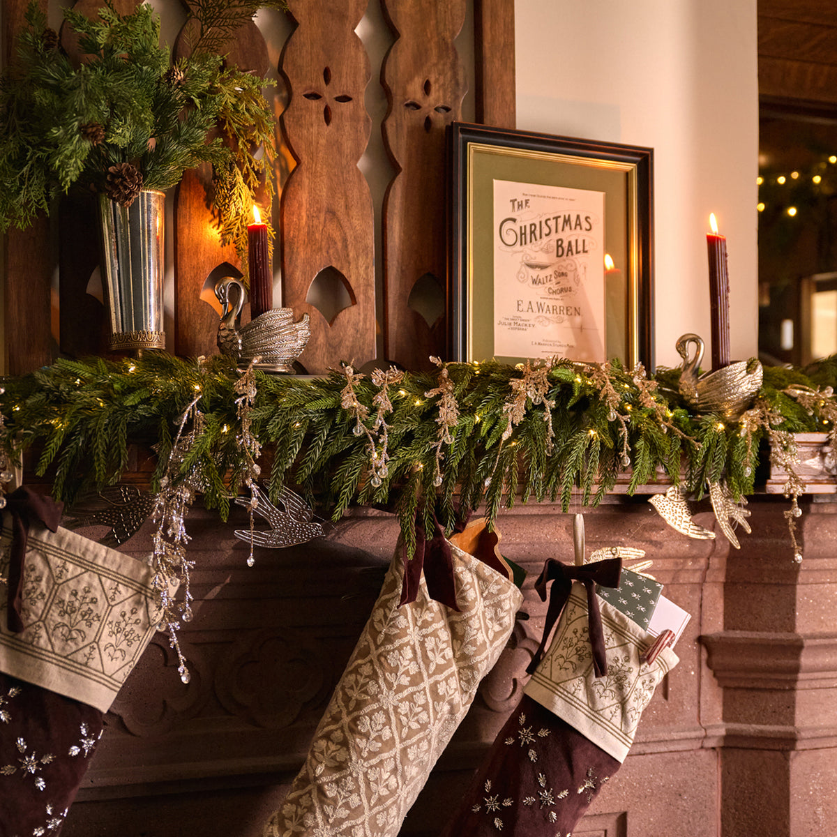 A festive holiday mantel decorated with green garlands, warm string lights, candles, and three patterned Christmas stockings hanging from a dark wood fireplace. A framed print reading 'The Christmas Ball' sits on the mantel.