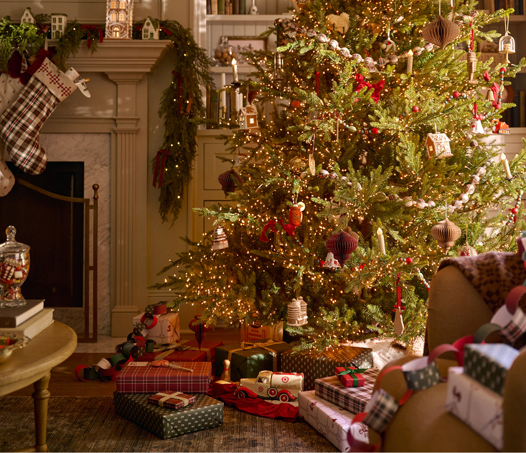 A beautifully decorated Christmas tree in a cozy living room, surrounded by wrapped gift boxes on the floor and stockings hanging by the fireplace.
