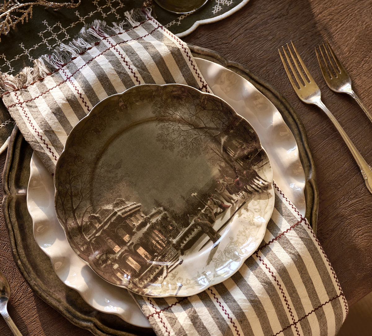 A place setting featuring a scalloped ceramic plate with a sepia-toned architectural landscape illustration, layered over a white charger plate and a striped beige and brown napkin, with two forks on a dark wood table.