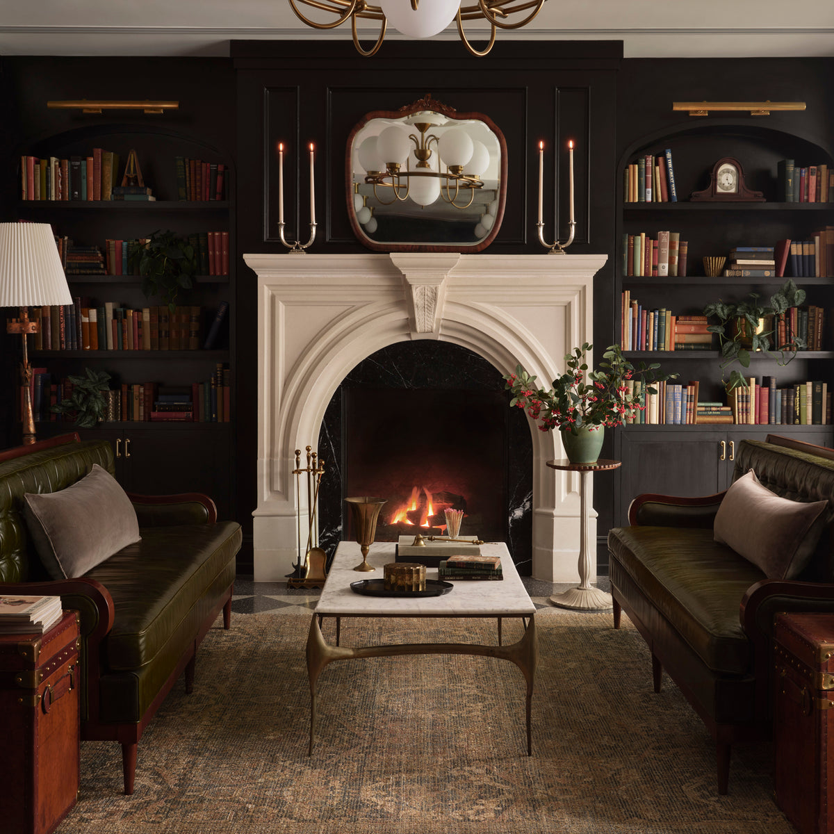 A classic dark, elegant living room featuring a white stone fireplace with a burning fire, flanked by tall bookshelves and two dark green leather sofas.