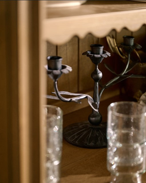 A dark metal three-arm candelabra with a branch-like design, shown in a warm indoor setting with blurred glassware in the foreground.