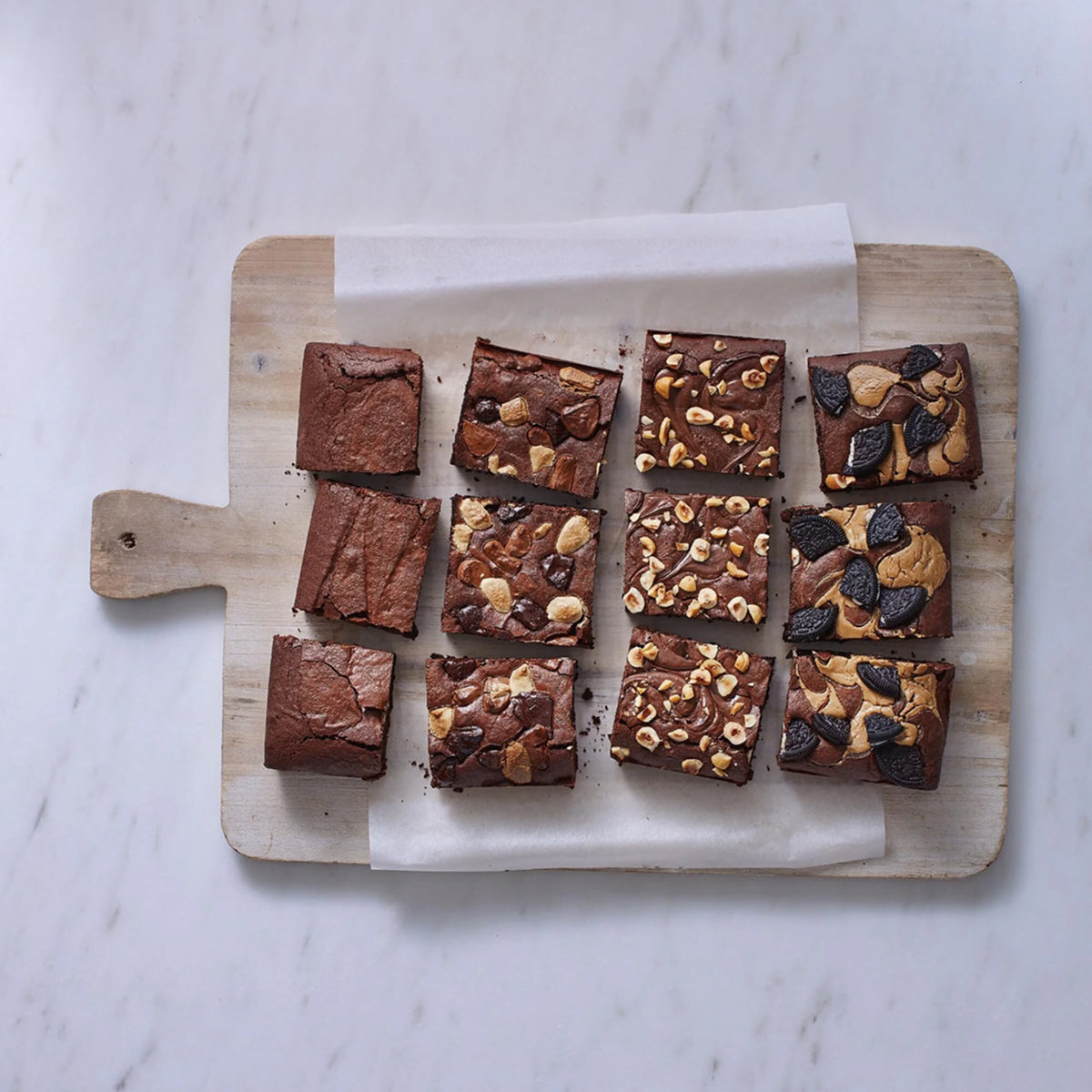 Assorted brownies on a wooden cutting board with white paper underneath on a marble surface.
