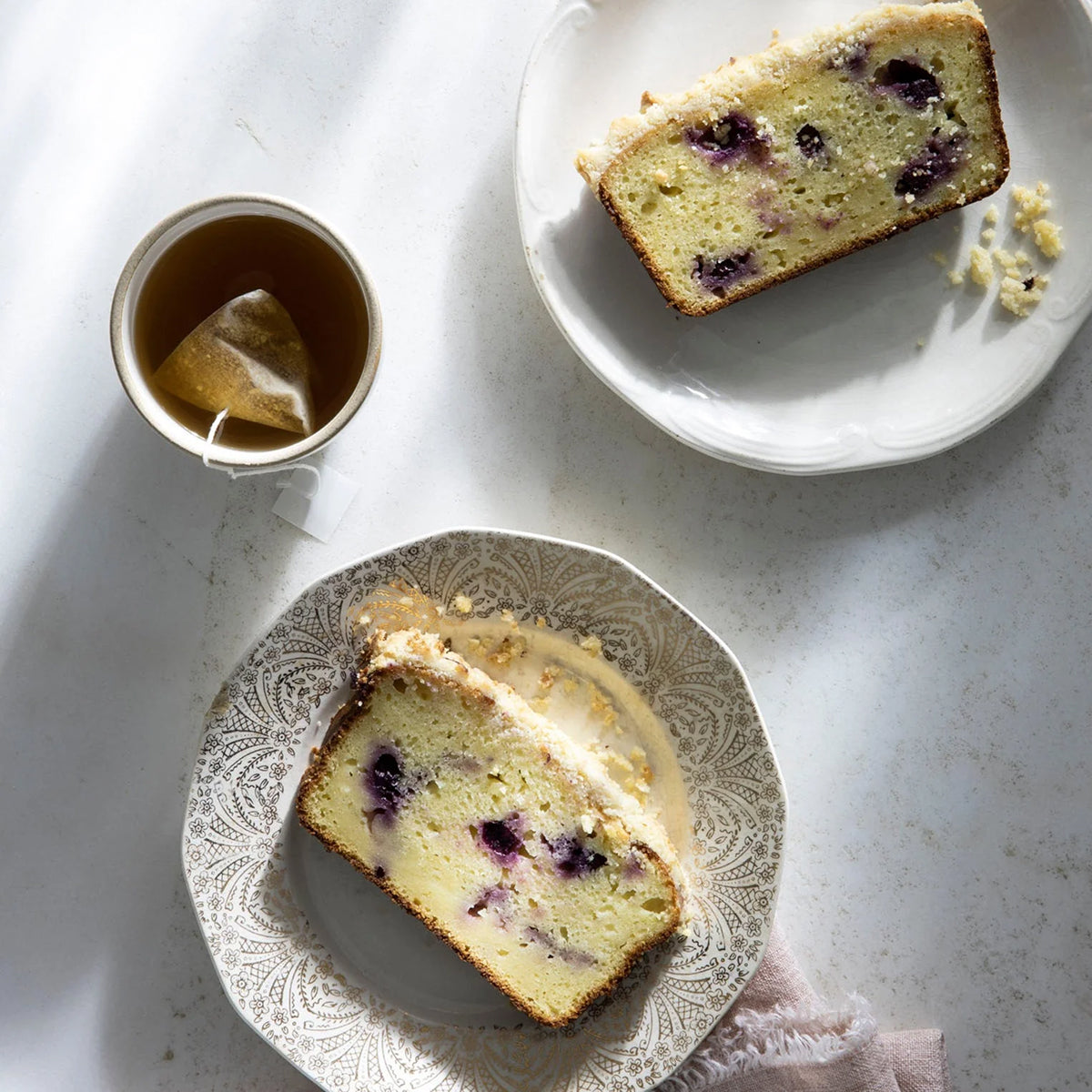 Two slices of lemon blueberry bread on decorative plates with a cup of tea on a light surface.