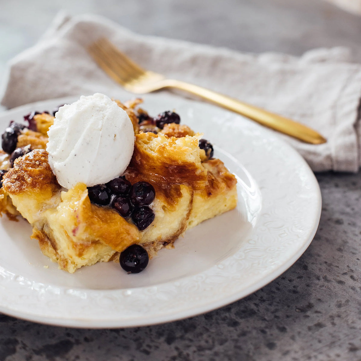 Slice of blueberry cake with ice cream on a white plate, gold fork beside, gray napkin.