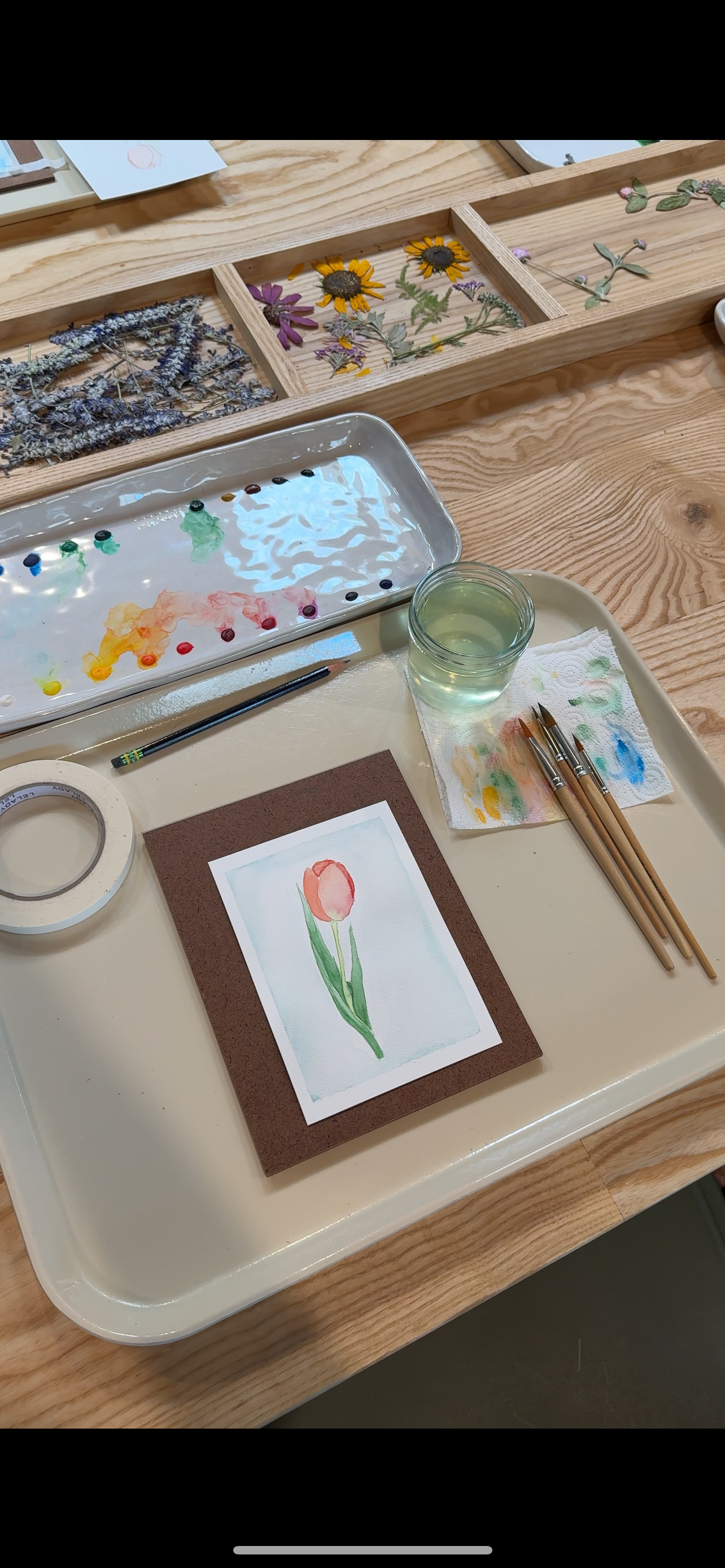 Artistic setup with watercolor paints, brushes, and a completed watercolor painting of a tulip on a wooden table.