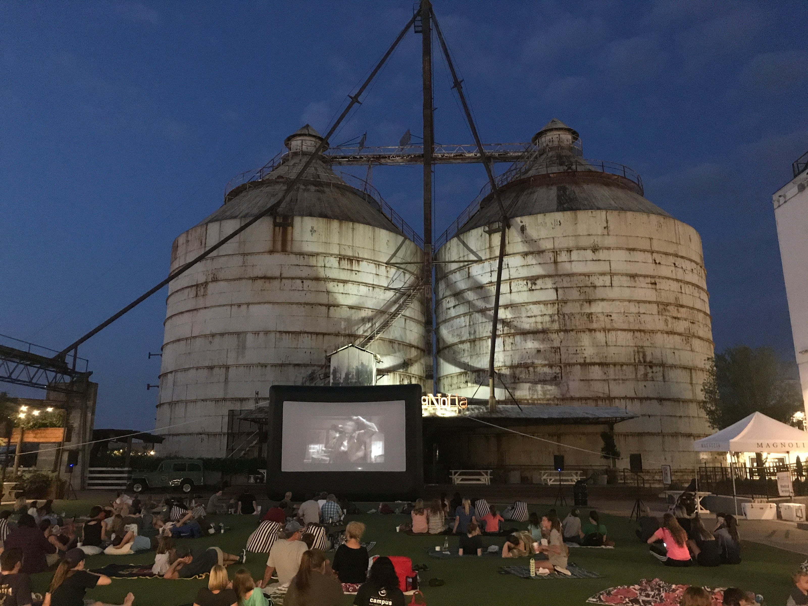 Silos with a movie screen projected onto them at night, with people gathered around.