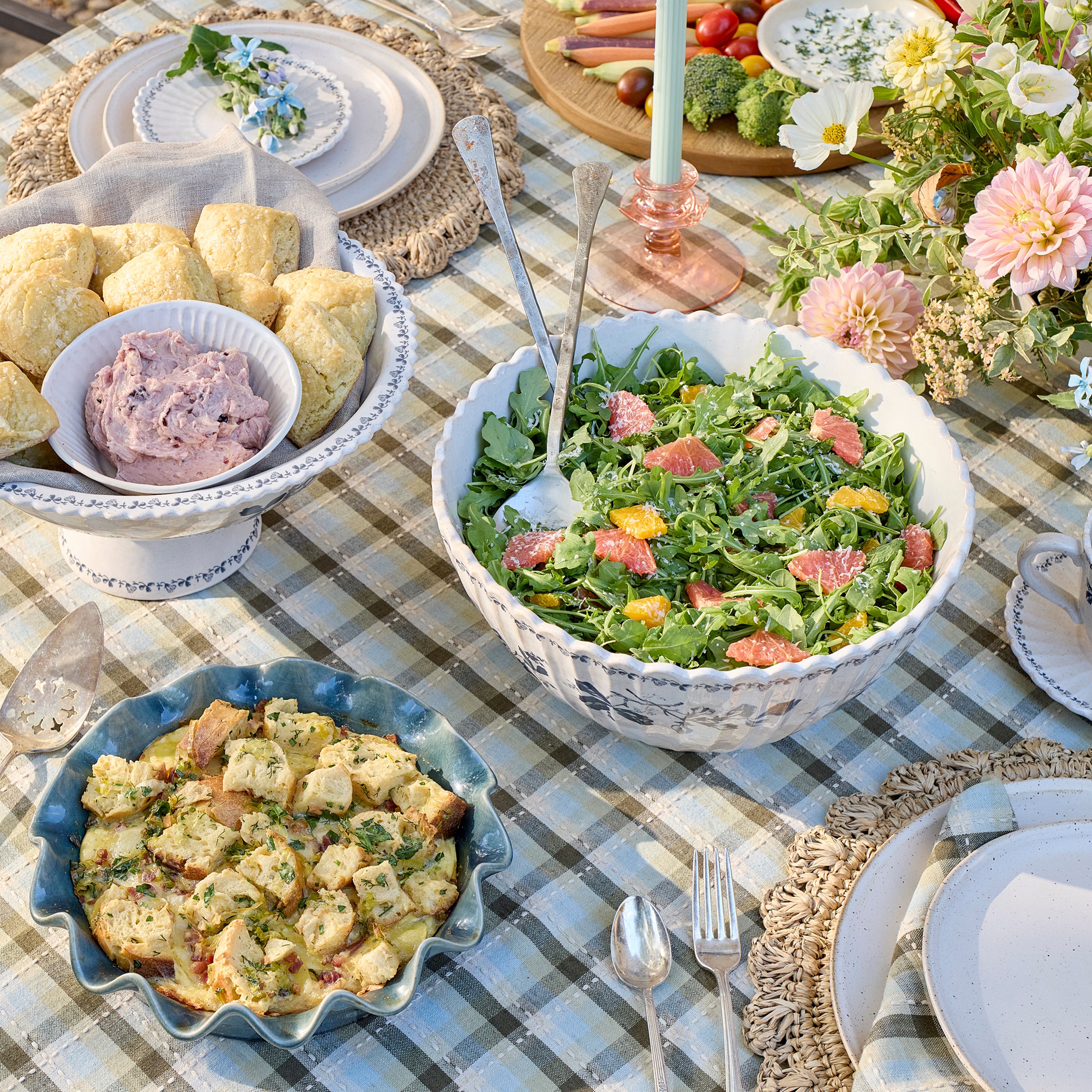 Outdoor table setting with a citrus mint salad, buttermilk biscuits, and potato leek strata and other dishes on a checkered tablecloth.