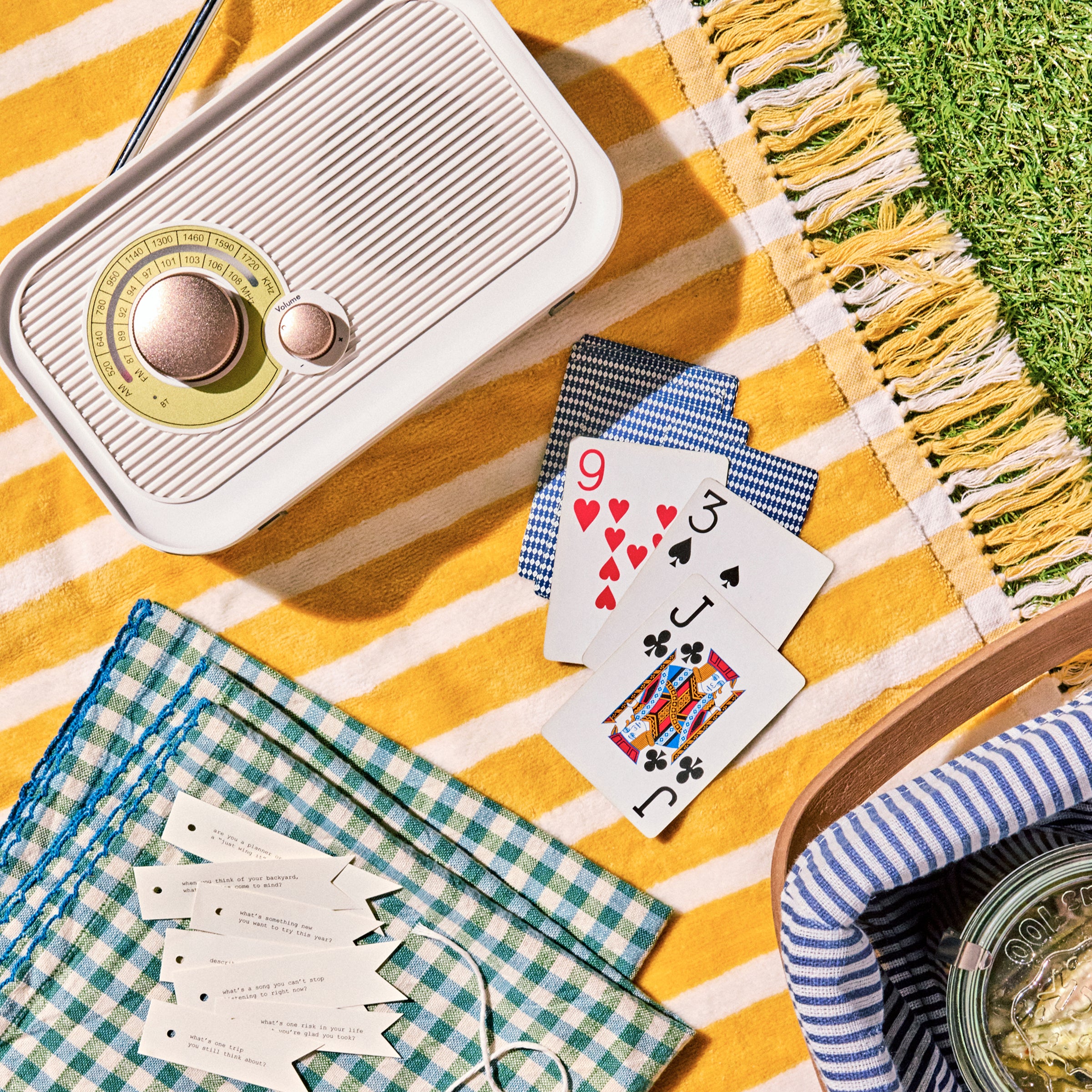 Vintage radio, playing cards, and checkered fabric on a yellow striped blanket outdoors.