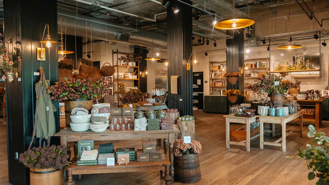 A rustic, cozy home decor and gift shop interior with wooden tables displaying ceramics, flowers, books, and home accessories under warm hanging pendant lights.