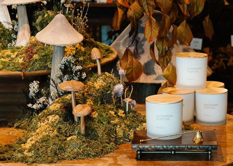 A styled product display featuring several white candles with light wood lids labeled 'GATHERED' on a wooden surface, alongside decorative faux moss, mushrooms, dried leaves, and small brass accessories.