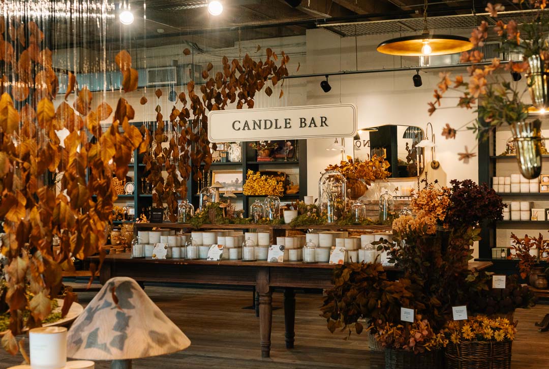A warm, rustic retail display for 'Candle Bar' featuring numerous white wax candles on a dark wooden table, surrounded by autumn foliage decorations and hanging warm lights in a cozy indoor setting.
