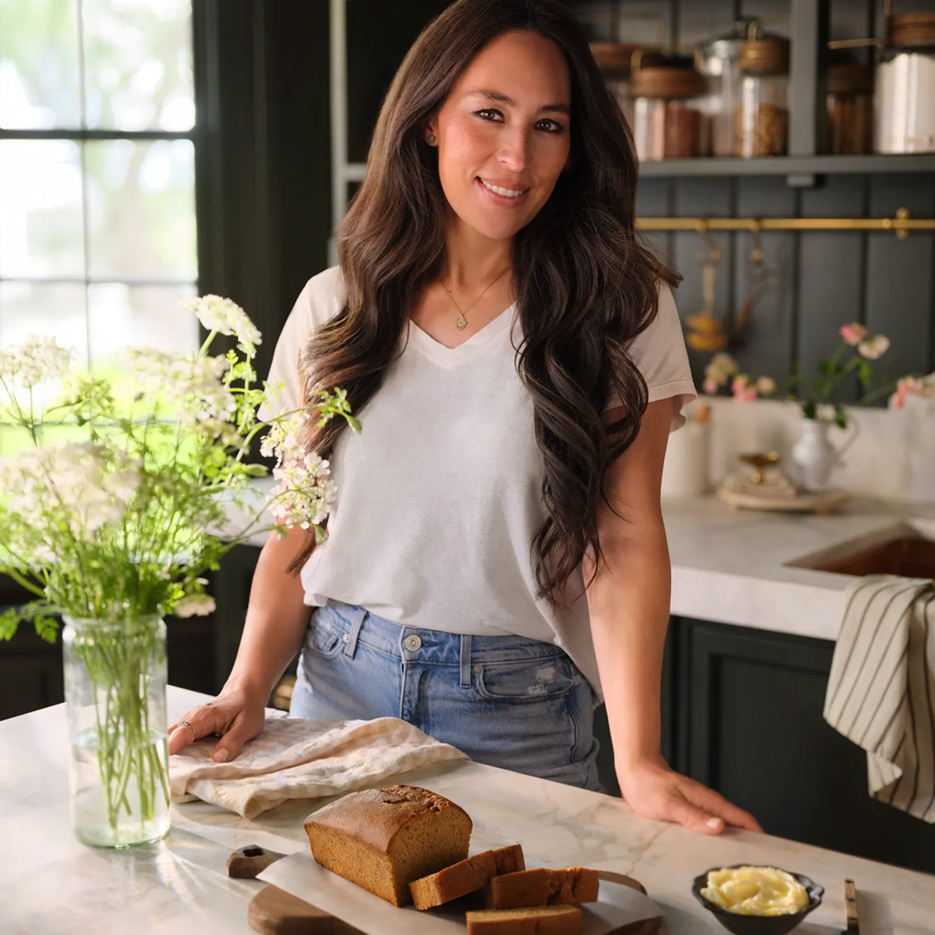 Joanna Gaines standing in her butler's pantry with a loaf of Magnolia Table After School Banana Bread on the kitchen island.