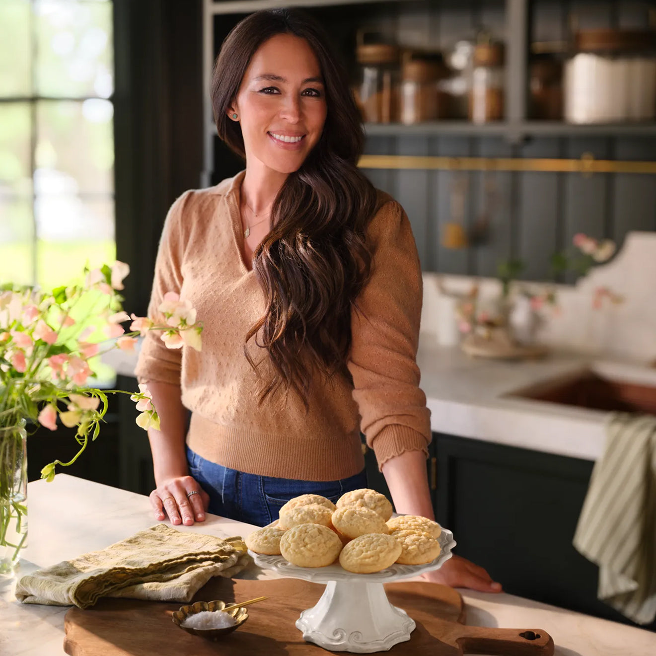 Joanna Gaines standing in her butler's pantry with Magnolia Table Classic Sugar Cookies on a cake stad on her kitchen island.