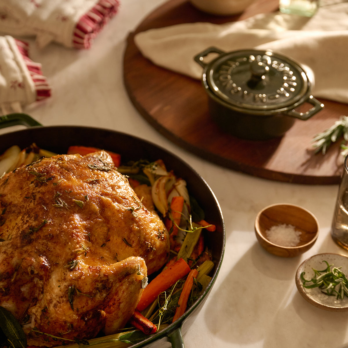 A roasted whole chicken with carrots and vegetables in a dark cast-iron skillet, styled on a dining table with a small lidded cast-iron pot on a wooden board, salt bowls, and a glass of water nearby.