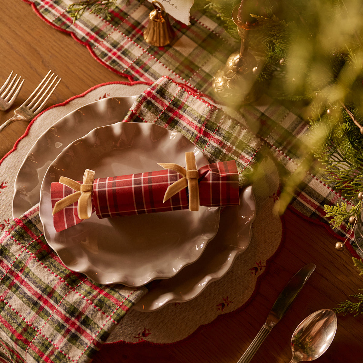 A styled holiday table setting with layered scalloped plates, a red and green plaid napkin tied with a beige bow, and a Christmas table runner on a wooden table.