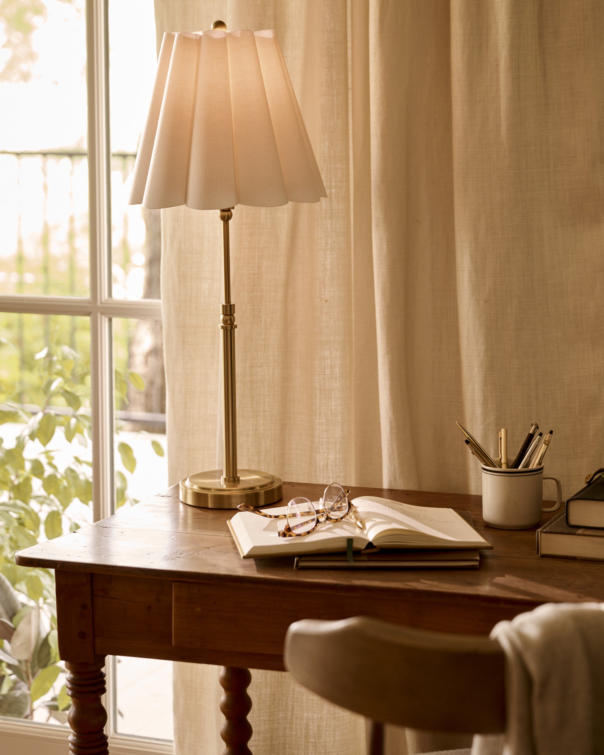 A lit table lamp with a pleated off-white shade and brass base sits on a wooden desk beside an open notebook, eyeglasses, and a mug with pens, in front of beige curtains by a window.