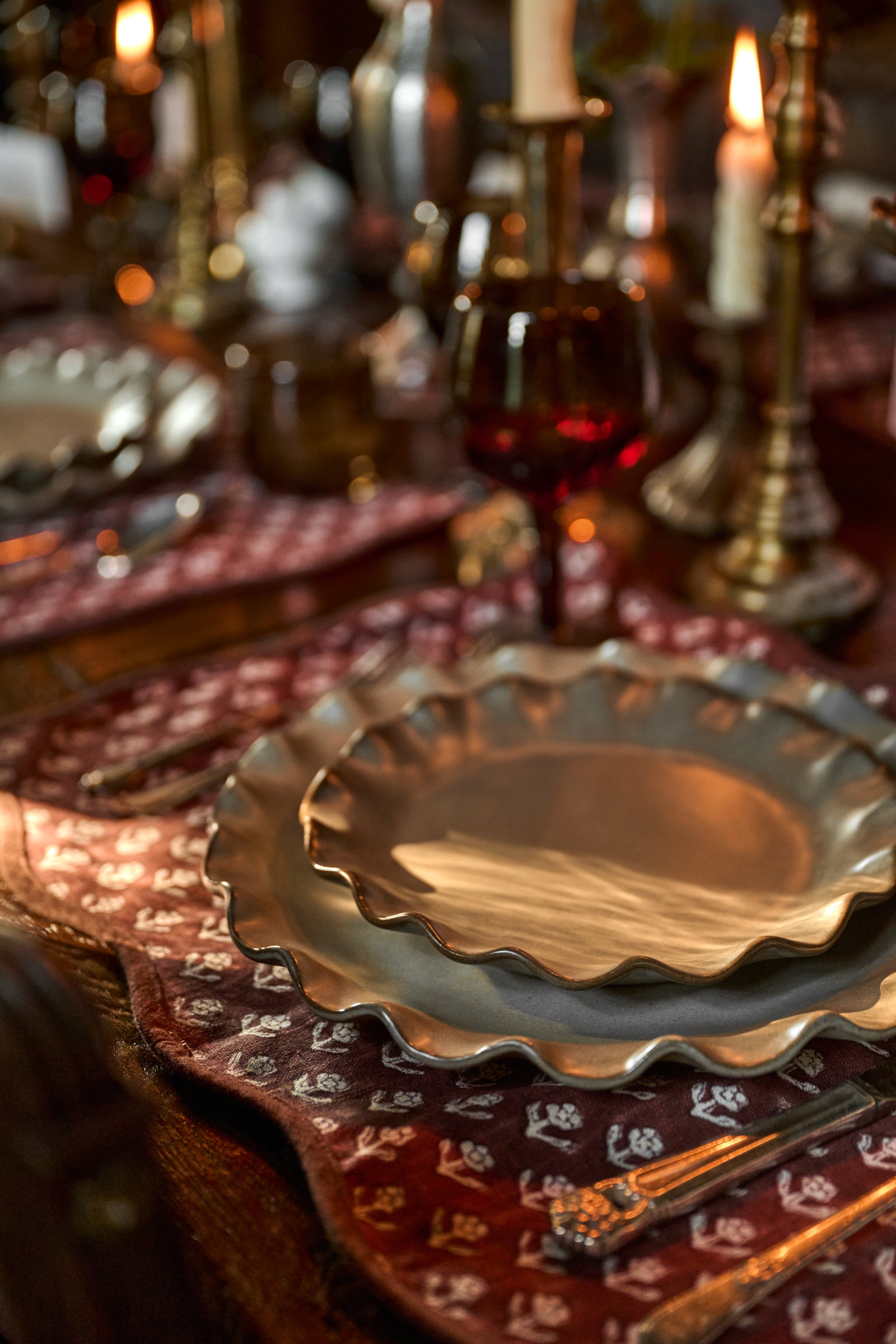 A close-up of a rustic table setting featuring a hammered silver charger plate on a patterned burgundy and gold placemat, with a candlelit background.