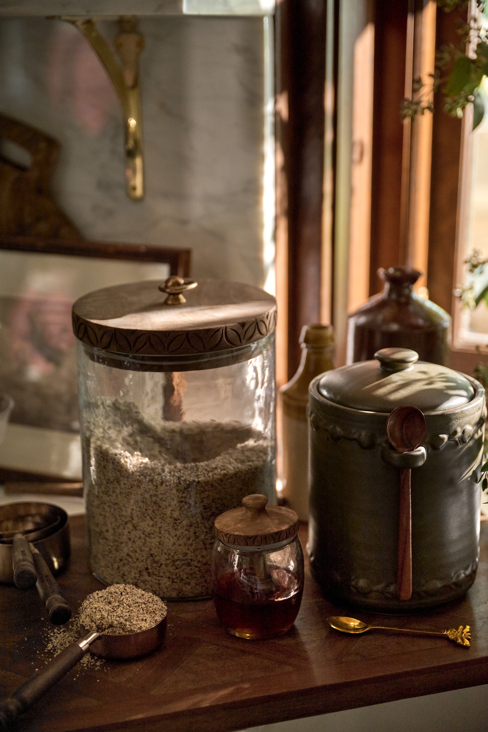 A rustic kitchen scene with glass and metal storage jars on a wooden table, including a large jar filled with oats or grains, a small jar of amber liquid, and a dark metal pot with a wooden spoon, set in warm natural light near a window.