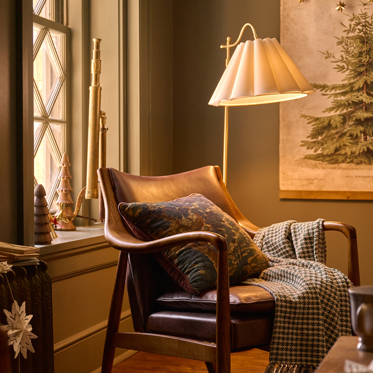 A warm, cozy interior scene featuring a mid-century modern leather armchair with a brown leather slipcover, styled with a patterned throw pillow, a houndstooth throw blanket, and a lit floor lamp next to a window.