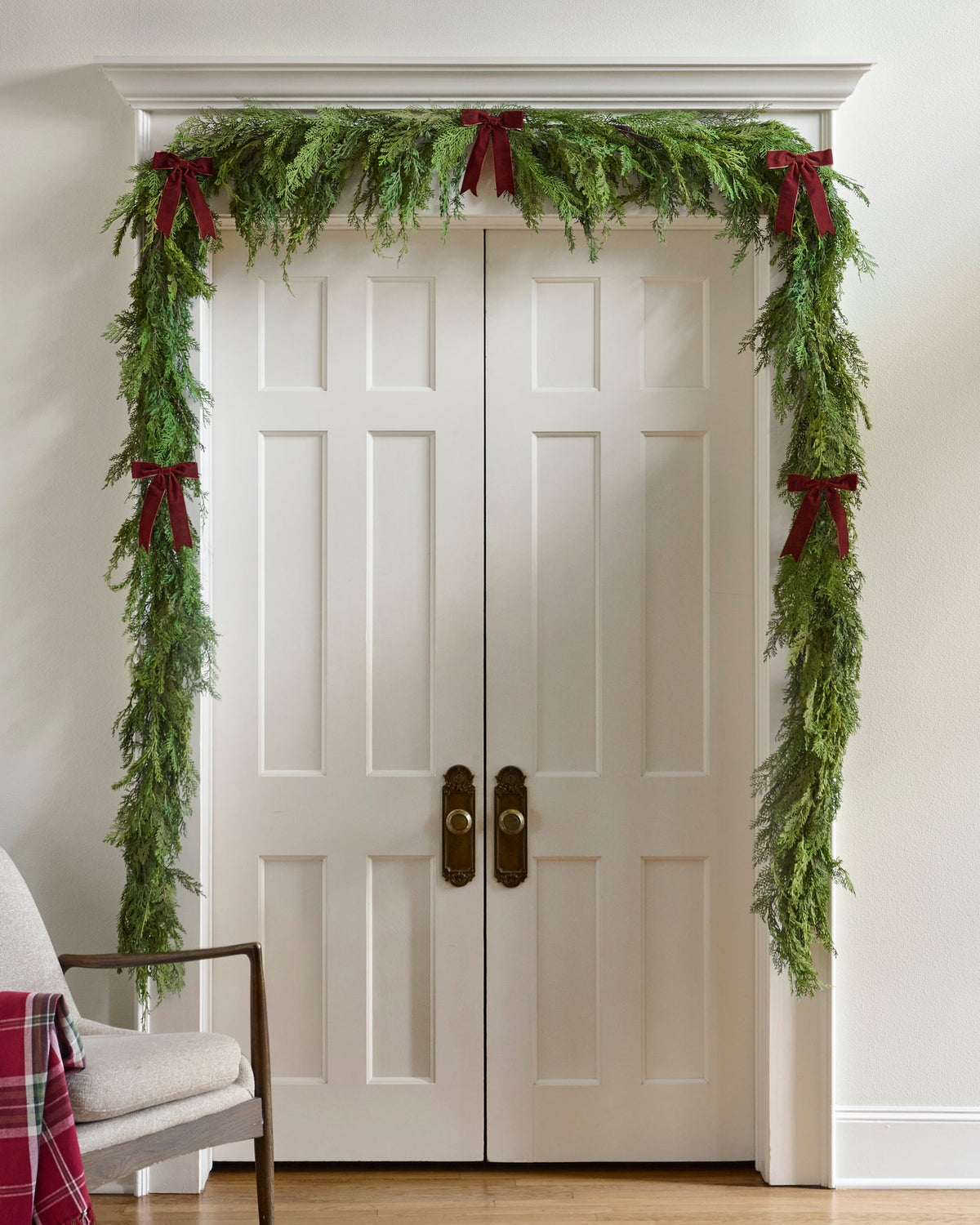 A green evergreen garland with dark red ribbon bows is draped over a pair of white double doors in a home entryway, with a chair partially visible in the lower left corner.