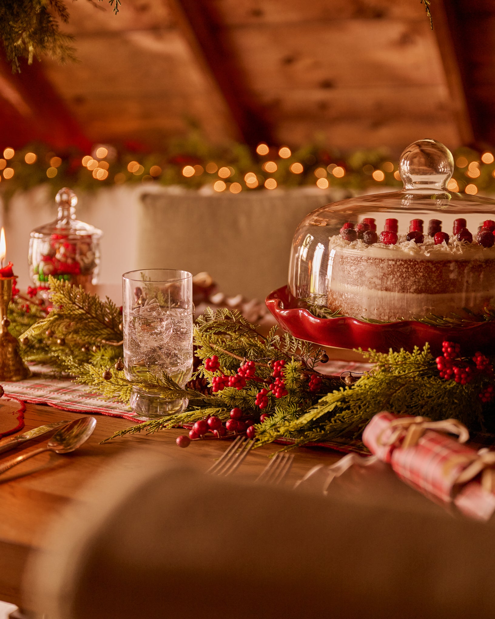 A festive holiday table setting with a tiered cake under a glass cloche, surrounded by green garlands, red berries, candles, and warm string lights in a cozy indoor environment.