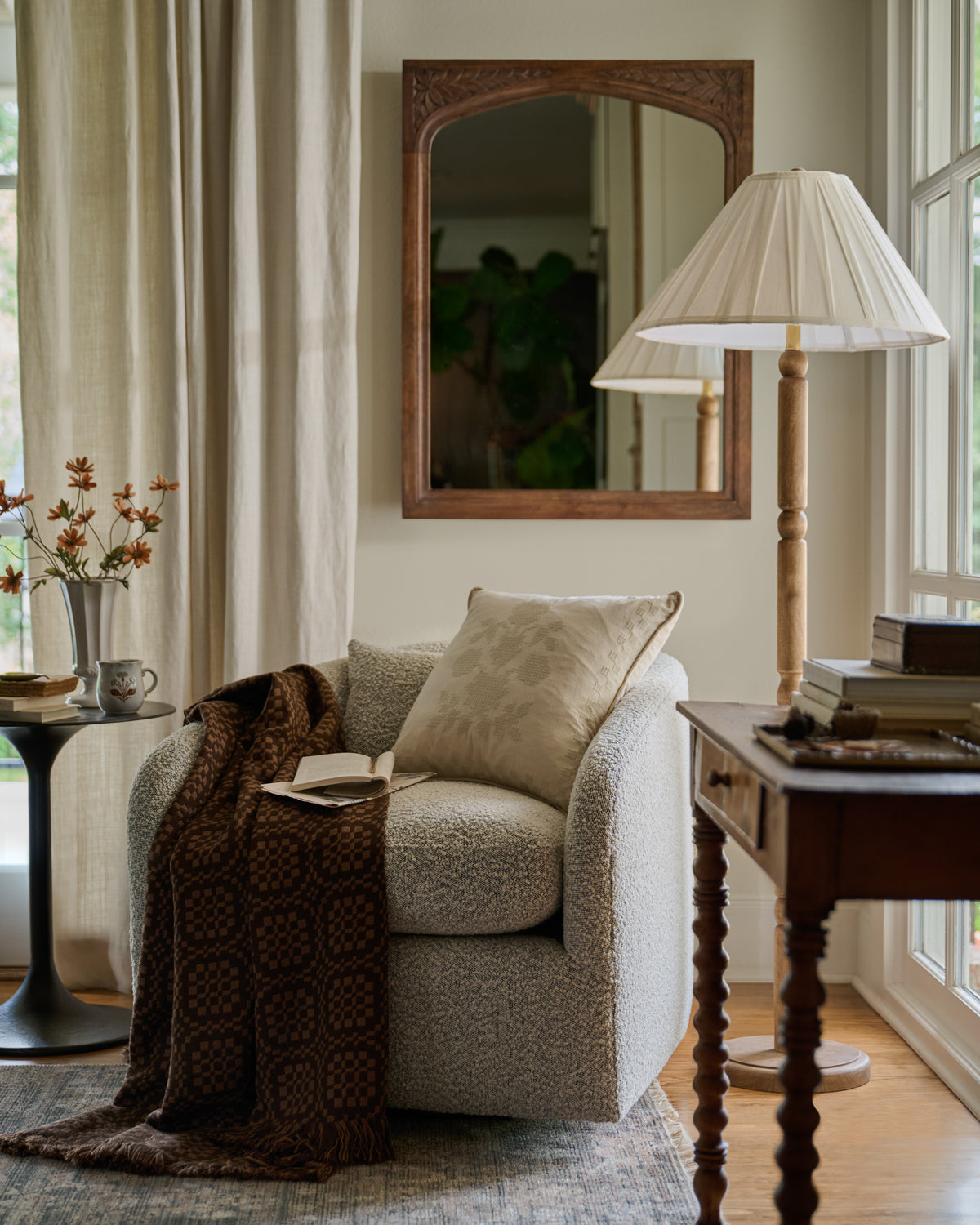 A cozy living room scene featuring a textured beige armchair with a matching pillow, draped with a dark brown patterned throw blanket. A tall floor lamp, a wooden side table with books, and a large wooden-framed mirror are arranged around the chair against cream walls and curtains.