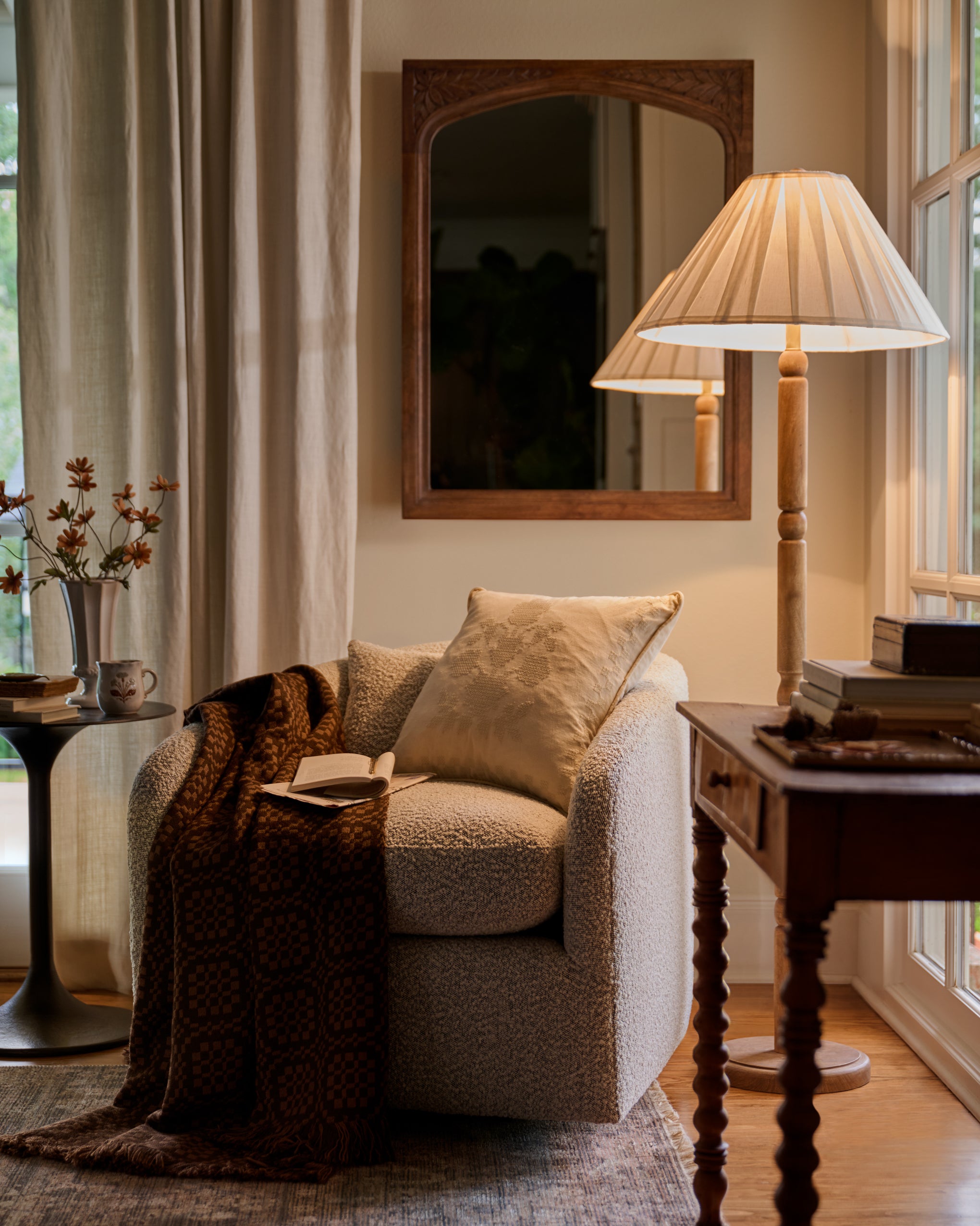 A cozy living room corner featuring a textured beige armchair with a matching pillow, draped with a dark brown patterned throw blanket. A tall floor lamp with a pleated cream shade stands on a wooden table next to the chair, and a wooden-framed mirror hangs on the wall above.