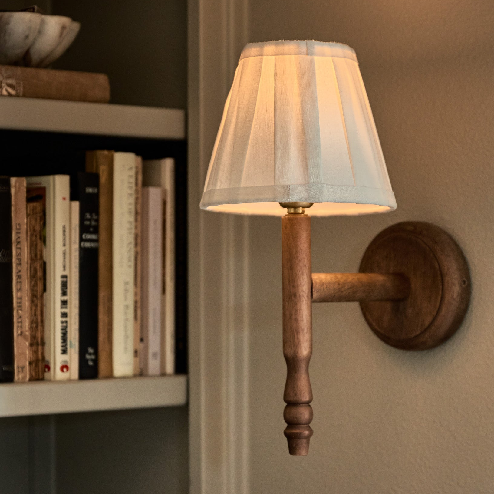 A rustic wall-mounted sconce with a white pleated fabric shade, turned wood arm, and round wooden backplate, shown lit against a beige wall next to a bookshelf.