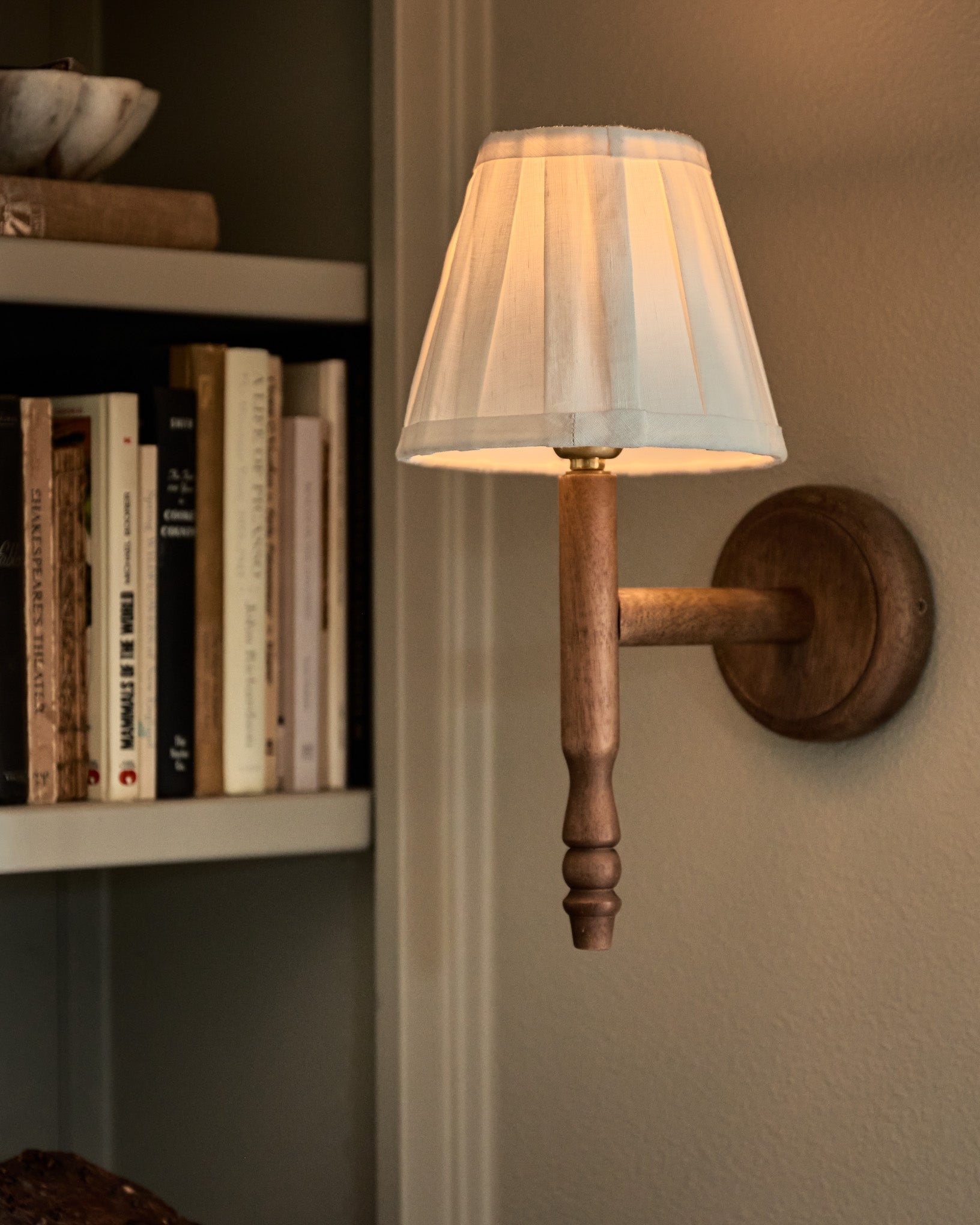 A wall-mounted sconce lamp with a white pleated fabric shade, a turned wood stem, and a round wooden wall plate, shown lit in a home interior next to a bookshelf.