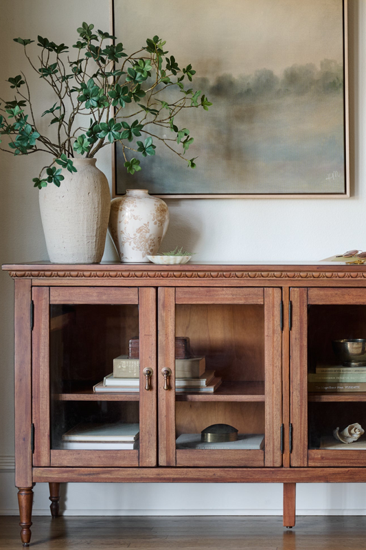 Wooden sideboard with glass doors displaying decorative items, vases, and a plant.