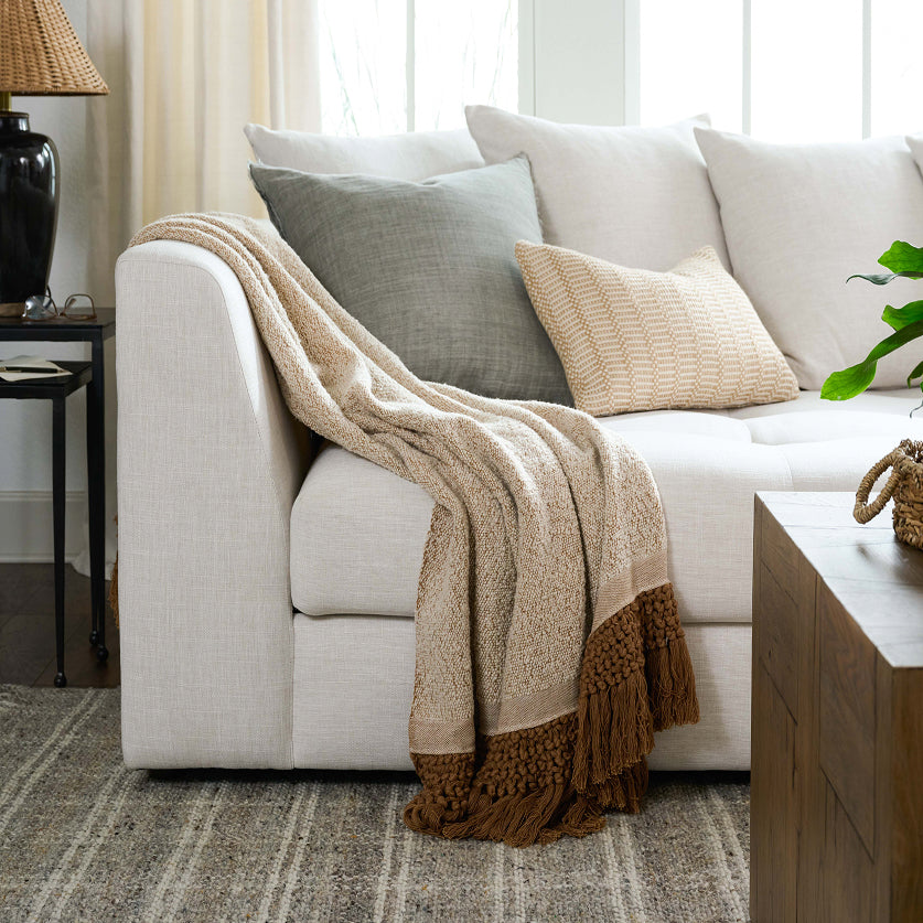 A cozy living room scene featuring a light beige textured throw blanket draped over a white sofa, styled with neutral-toned pillows and a striped area rug.
