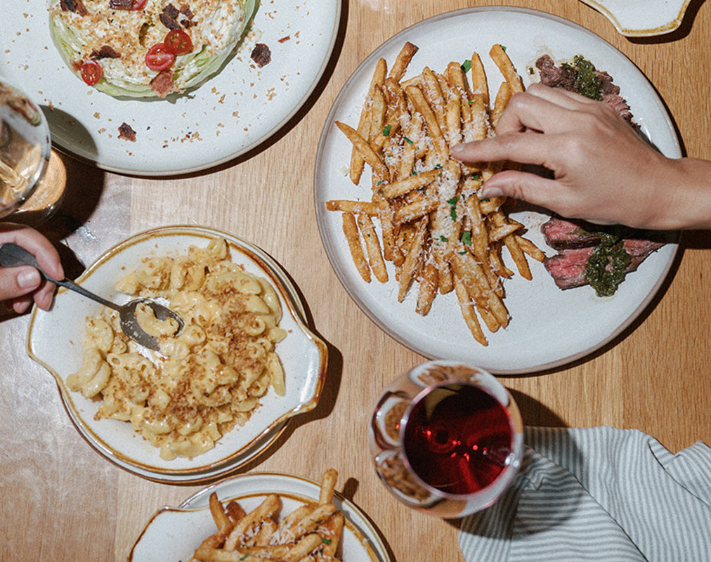Diverse food dishes on a table with a hand reaching for fries. Book your holiday gathering.  Celebrate meaningful moments this season with an event at Magnolia Table.  Inquire here.