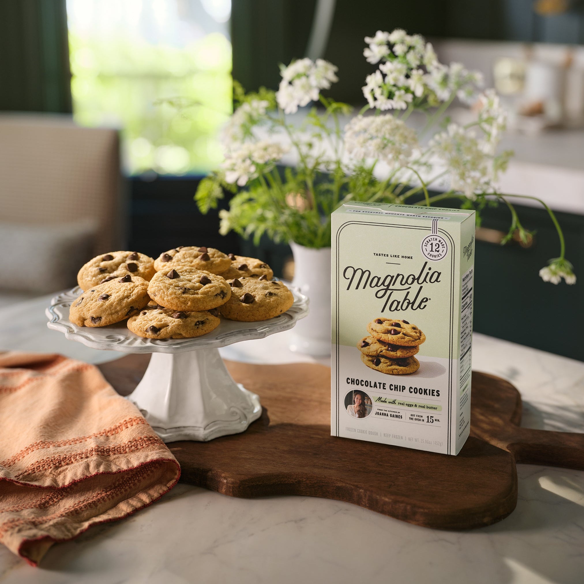 Box of Magnolia Table Frozen Chocolate Chip Cookie Dough on a kitchen island with Magnolia Table Chocolate Chip Cookies on a plate stand and flowers in the background.