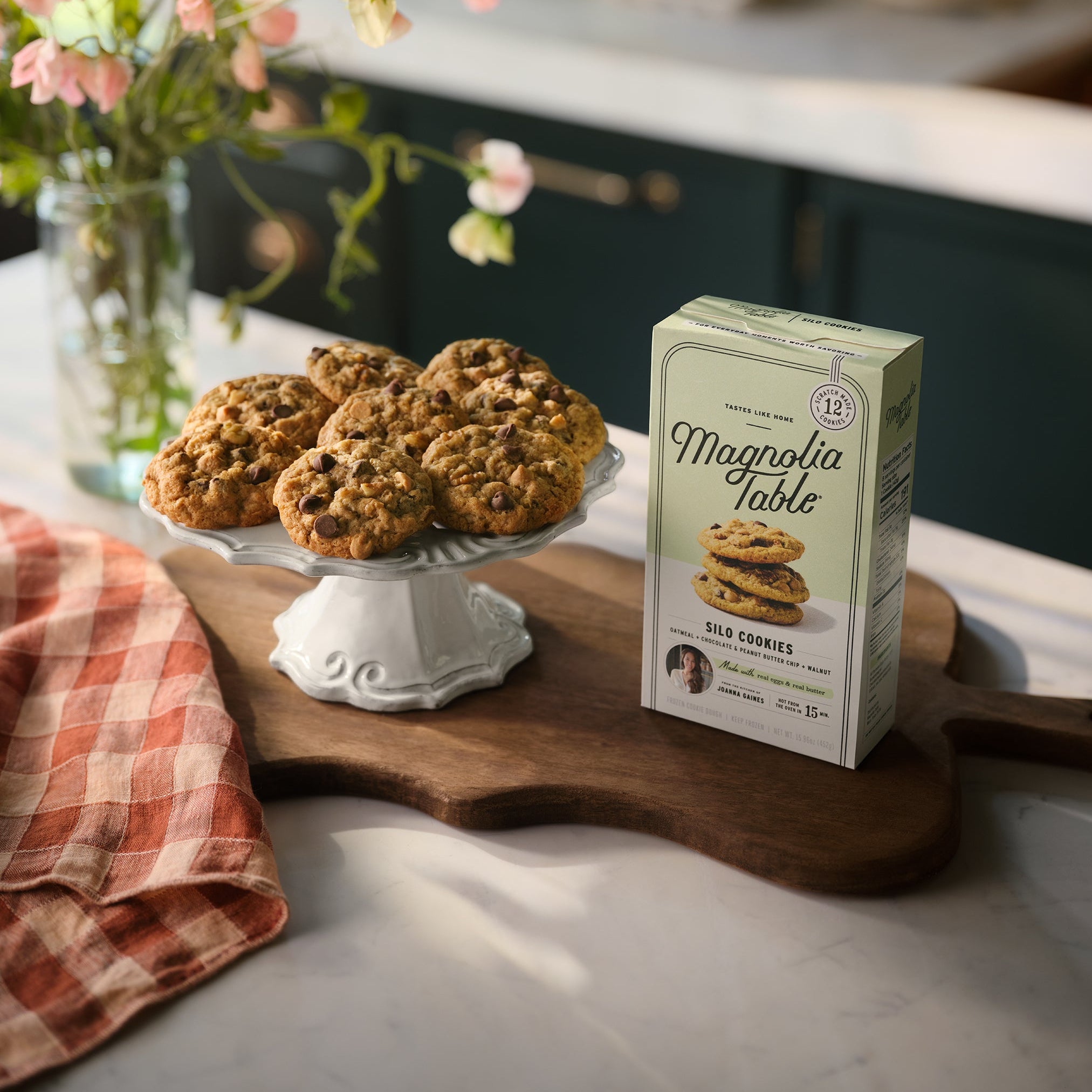 Box of Magnolia Table Frozen Silo Cookie Dough on a kitchen island with Magnolia Table Silo Cookies on a plate stand and flowers in the background.