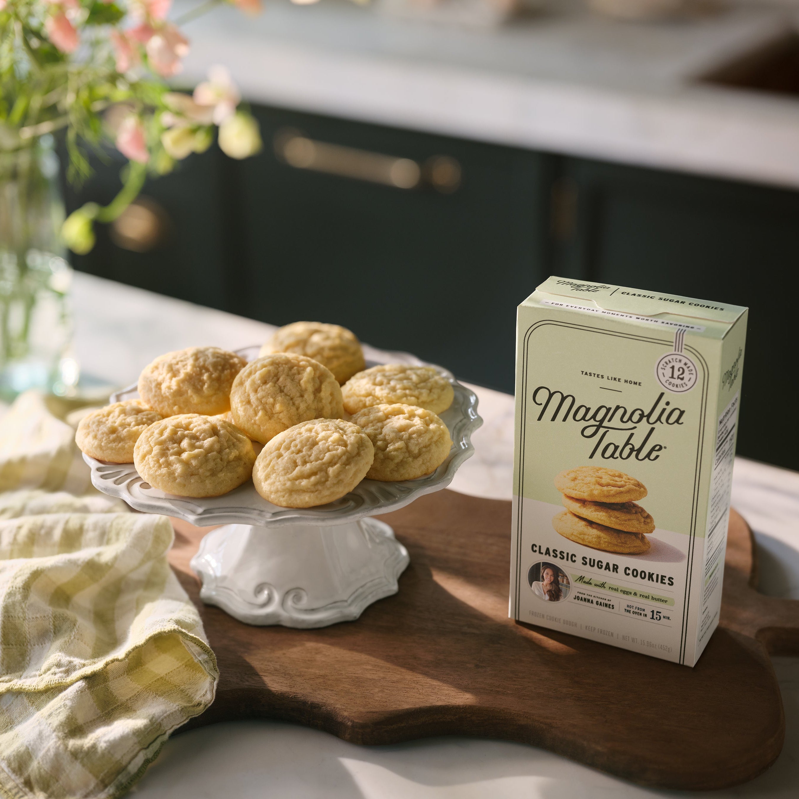 Box of Magnolia Table Frozen Classic Sugar Cookie Dough on a kitchen island with Magnolia Table Classic Sugar Cookies on a plate stand and flowers in the background.