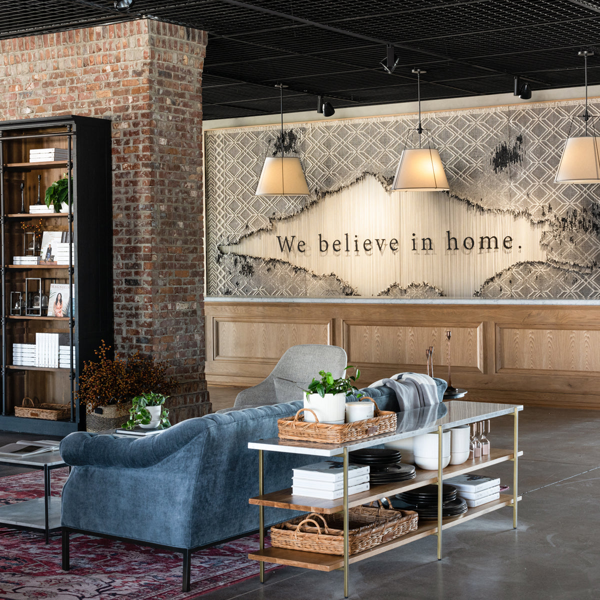 A styled interior room scene with a blue velvet sofa, a glass-top coffee table, brick wall, wood paneling, pendant lights, and a large wall mural that reads 'We believe in home.'.