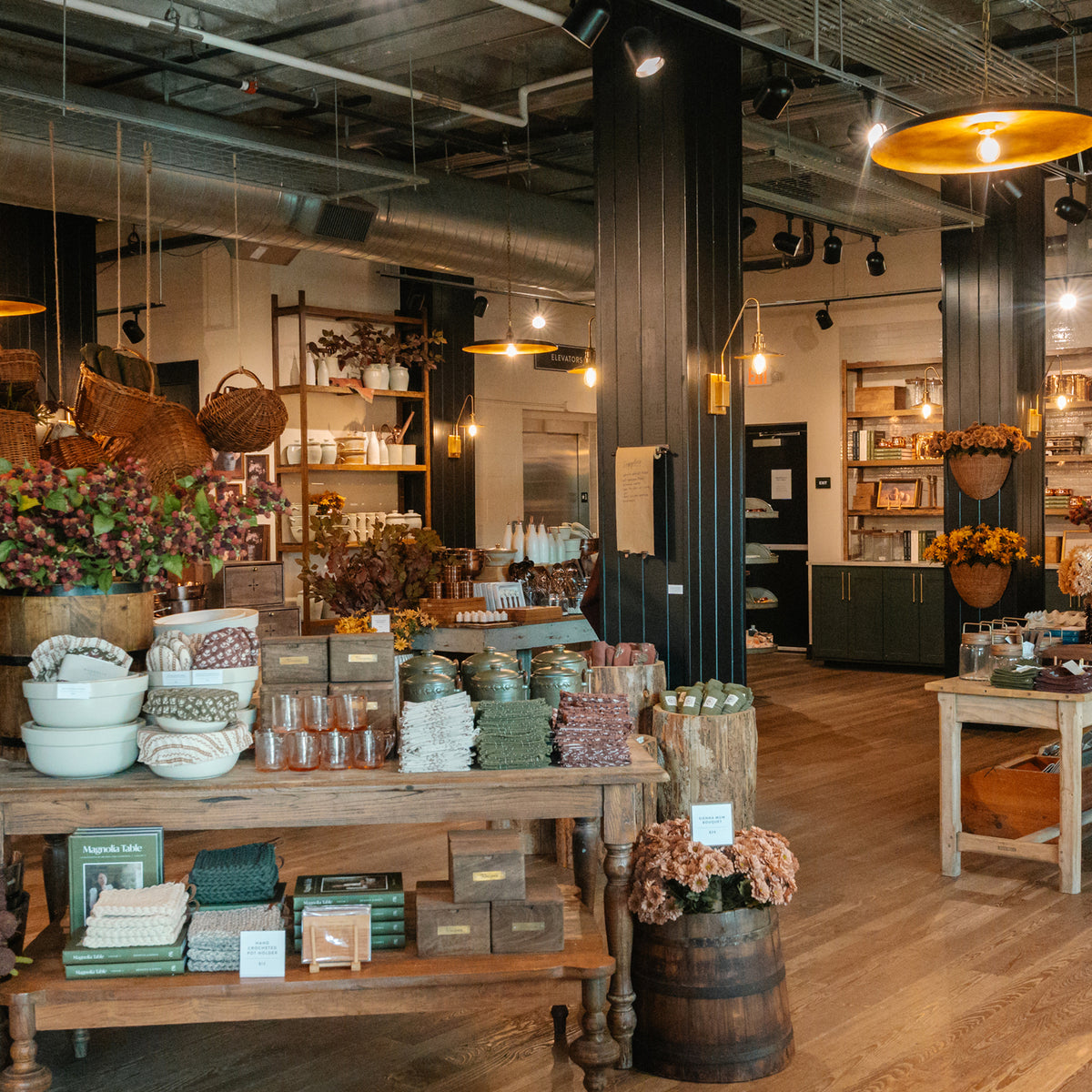 A rustic home goods store interior with wooden display tables, shelves, hanging baskets, and warm pendant lights. The scene shows assorted decorative items, textiles, flowers, and packaged goods arranged in a real retail environment.
