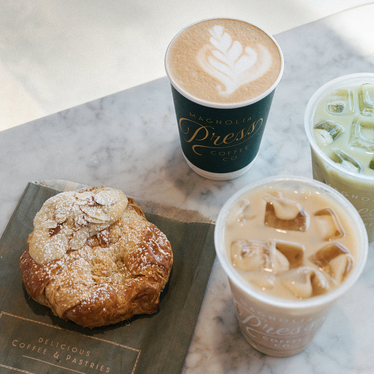A styled coffee and pastry flat-lay on a light marble surface. The set includes a croissant dusted with powdered sugar on a branded Magnolia Press paper bag, a tall latte with leaf latte art in a dark green Magnolia Press coffee cup, an iced coffee in a clear plastic cup, and a green iced tea in another clear cup.