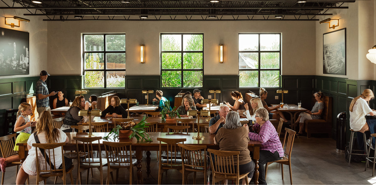 A large indoor study or library space with rows of wooden tables and chairs, many occupied by people working at desk lamps. Tall windows with dark frames line the back wall, and greenery is visible outside.